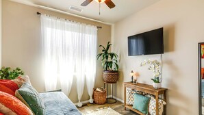 A well-lit living room with a ceiling fan, a large window with sheer curtains, a flat-screen TV mounted on the wall, a wooden console table with decorative items, and a cozy sofa with patterned cushions.