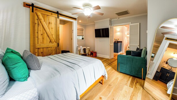 Een slaapkamer met een bed, een houten deur, een plafondventilator, een spiegel en een zichtbare badkamer door een open deur.