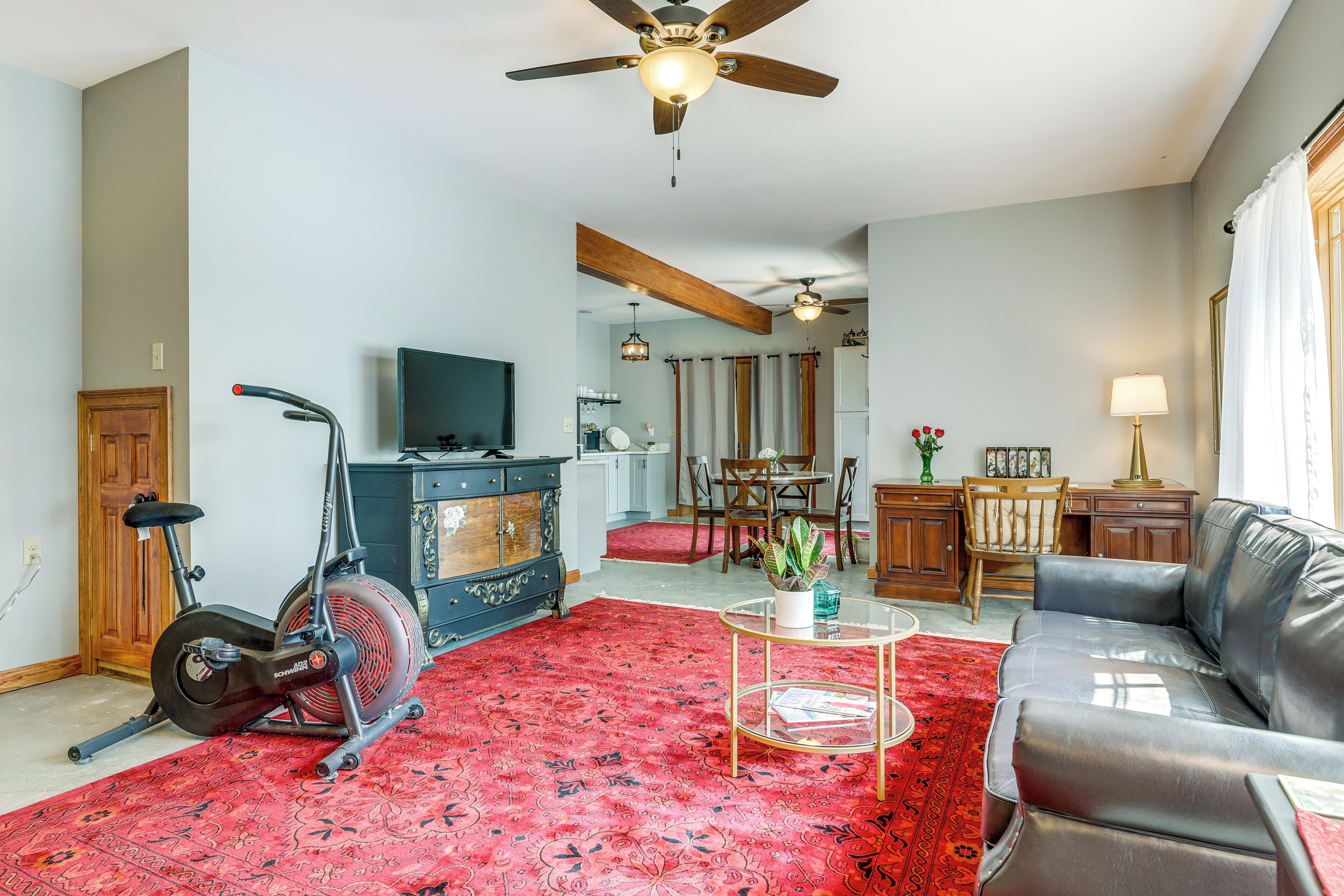 A living room with a red patterned carpet, a black leather sofa, a ceiling fan, a treadmill, and a television on a wooden cabinet.
