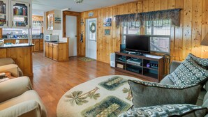 A cozy cabin interior with wooden walls, a kitchen area, a living room with a sofa and a television, and a ceiling fan. - Satsuma Home w/ Fire Pit + Dunns Creek Access! (Satsuma)