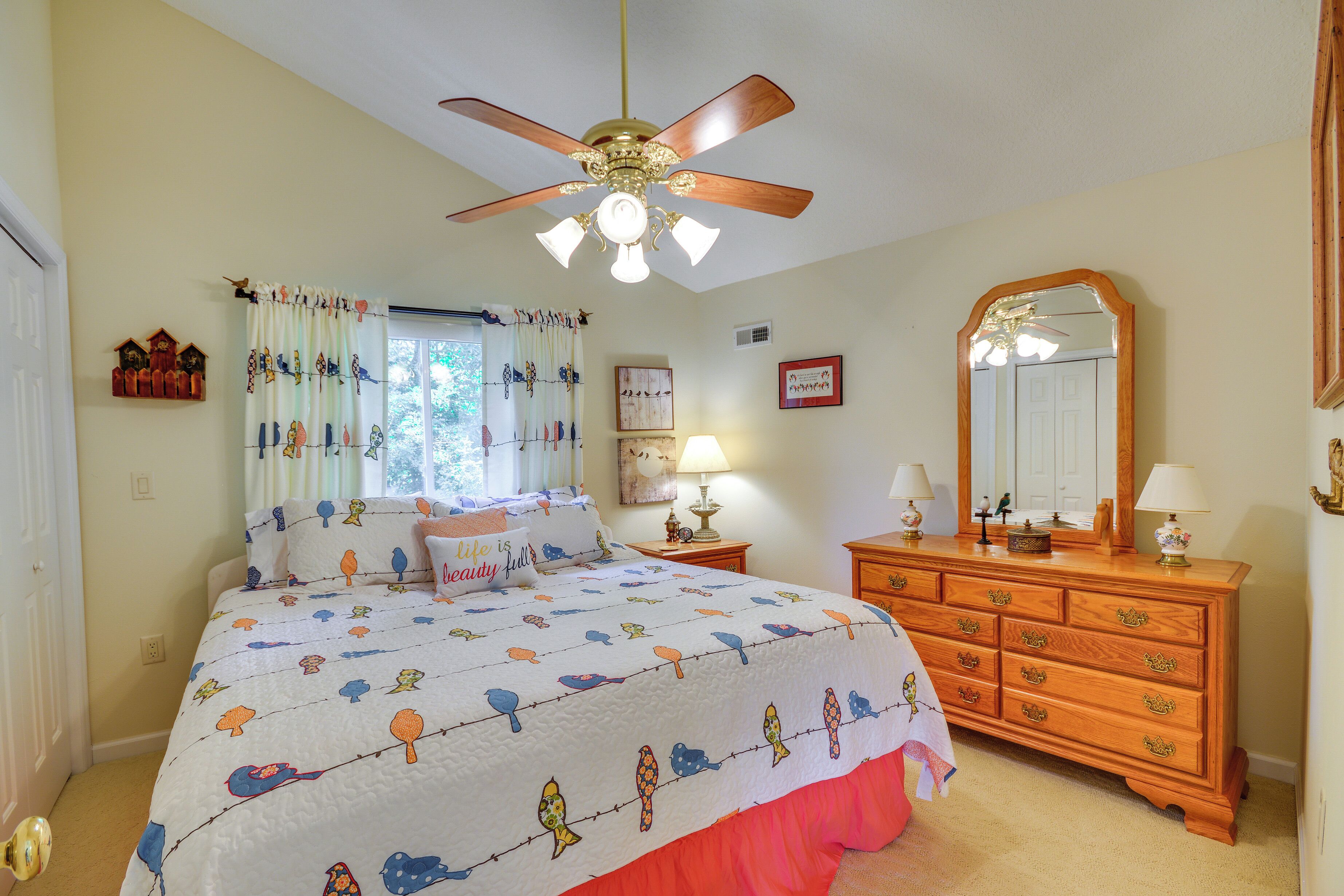 A bedroom with a bed, a dresser, a ceiling fan, a window with curtains, and wall decorations.