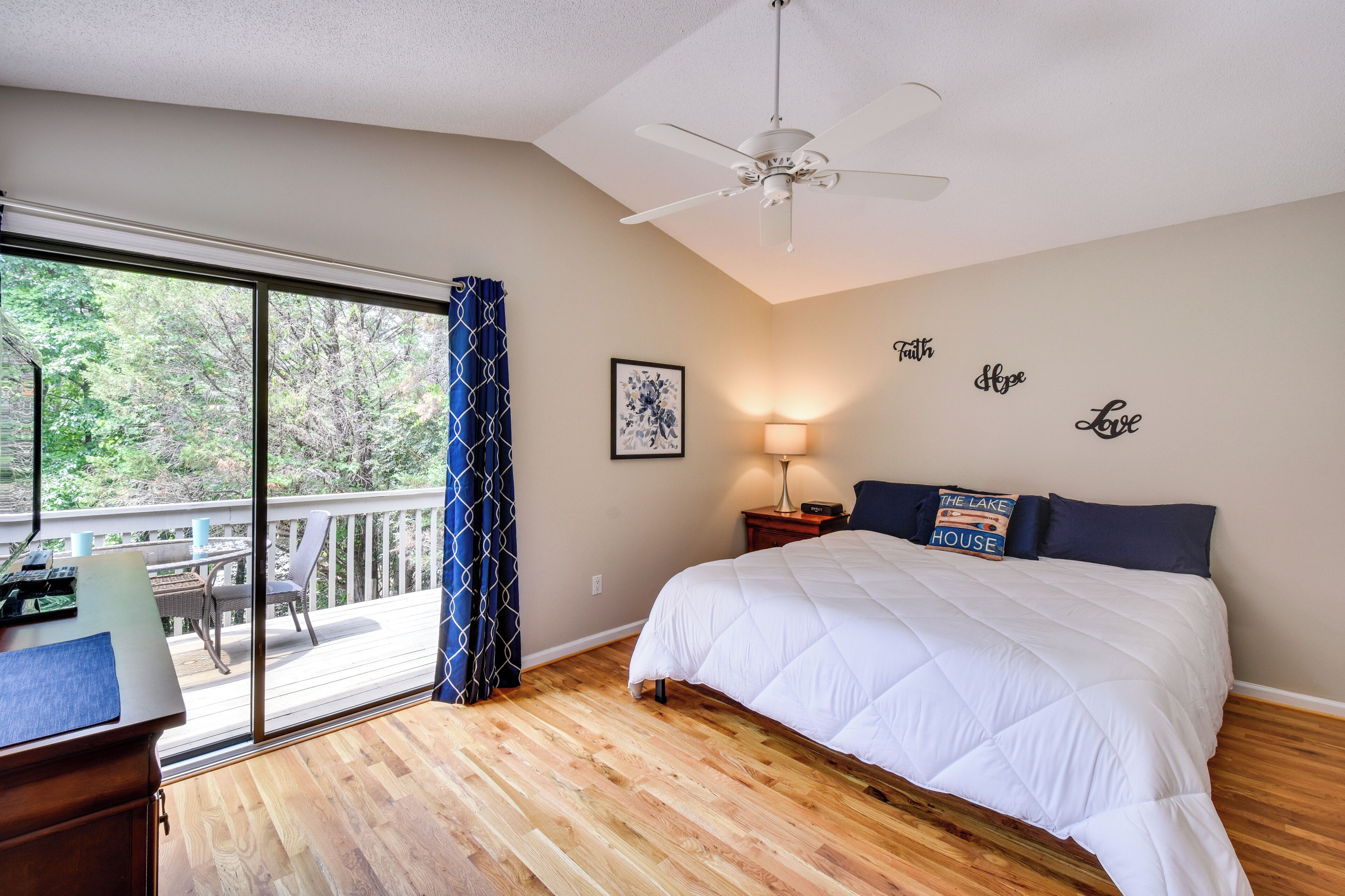 A bedroom with a large bed, a ceiling fan, a desk with a chair, a window with curtains, and a view of the outdoors.