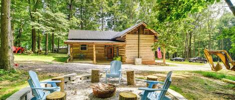 Eine rustikale Holzhütte mit einem Metalldach, umgeben von einem Wald. Im Vordergrund befinden sich zwei Adirondack-Stühle und eine Feuerstelle.
