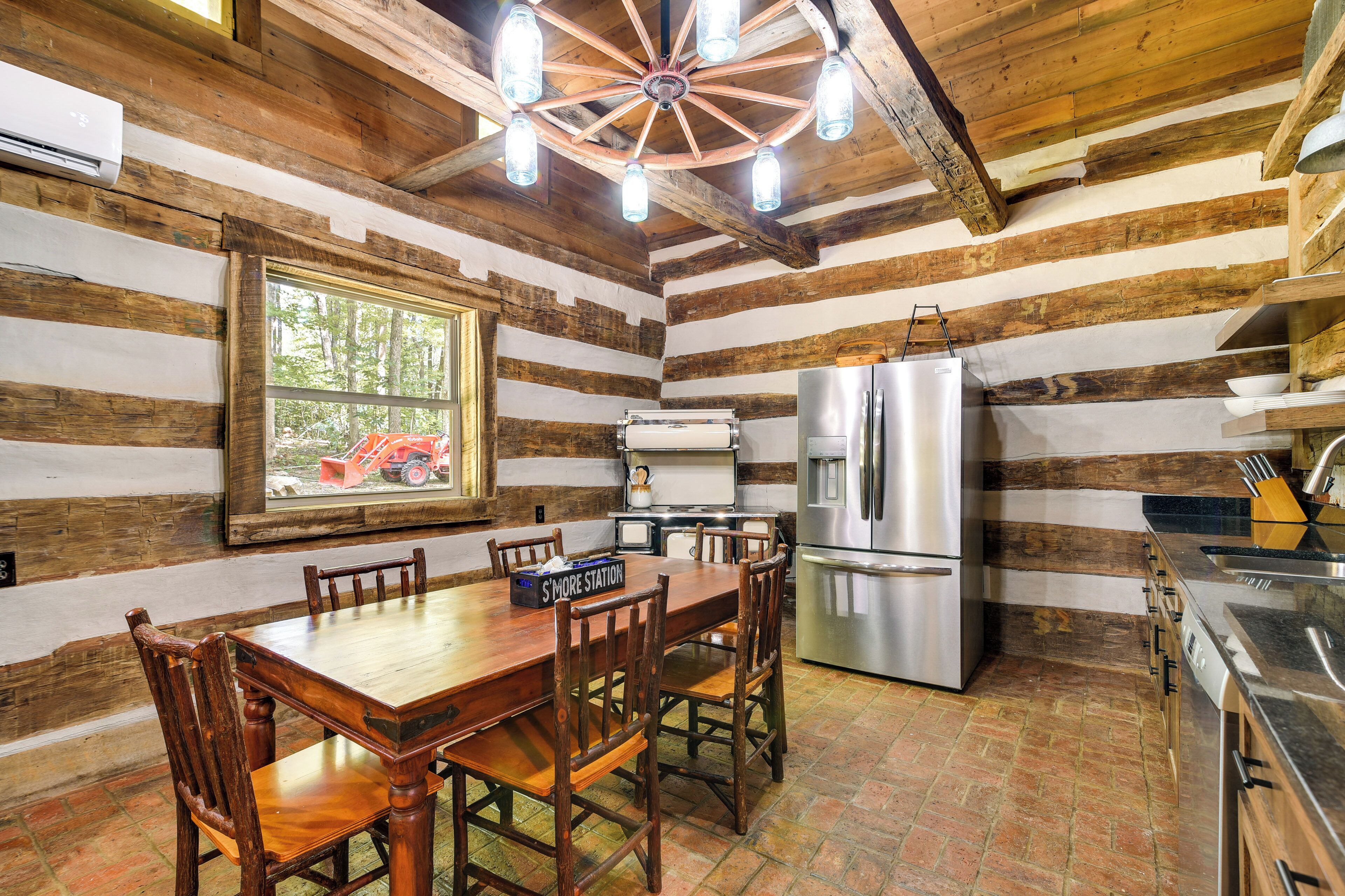 A rustic cabin interior with a wooden dining table, chairs, a refrigerator, and a window with a view of a tractor.