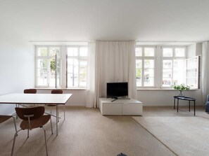 Een kamer met een witte tafel, stoelen, een televisie op een wit kastje en grote ramen met gordijnen.