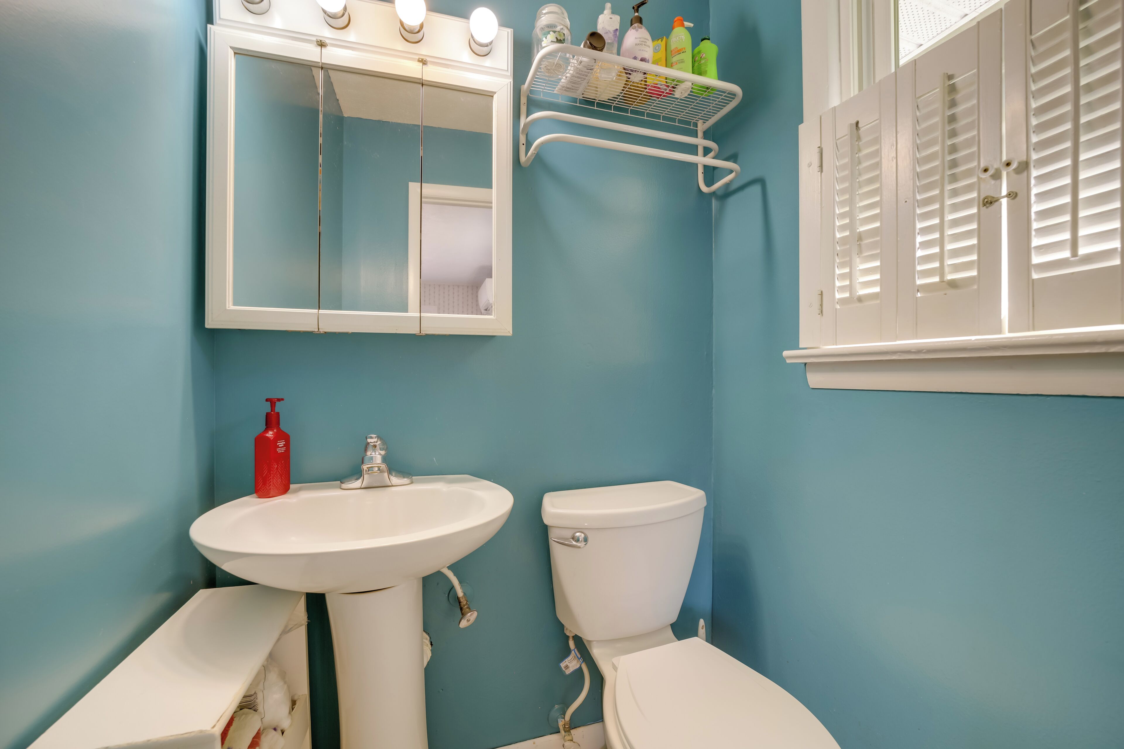 A bathroom with a white sink, a toilet, and a mirror. There is a shelf with toiletries above the mirror.