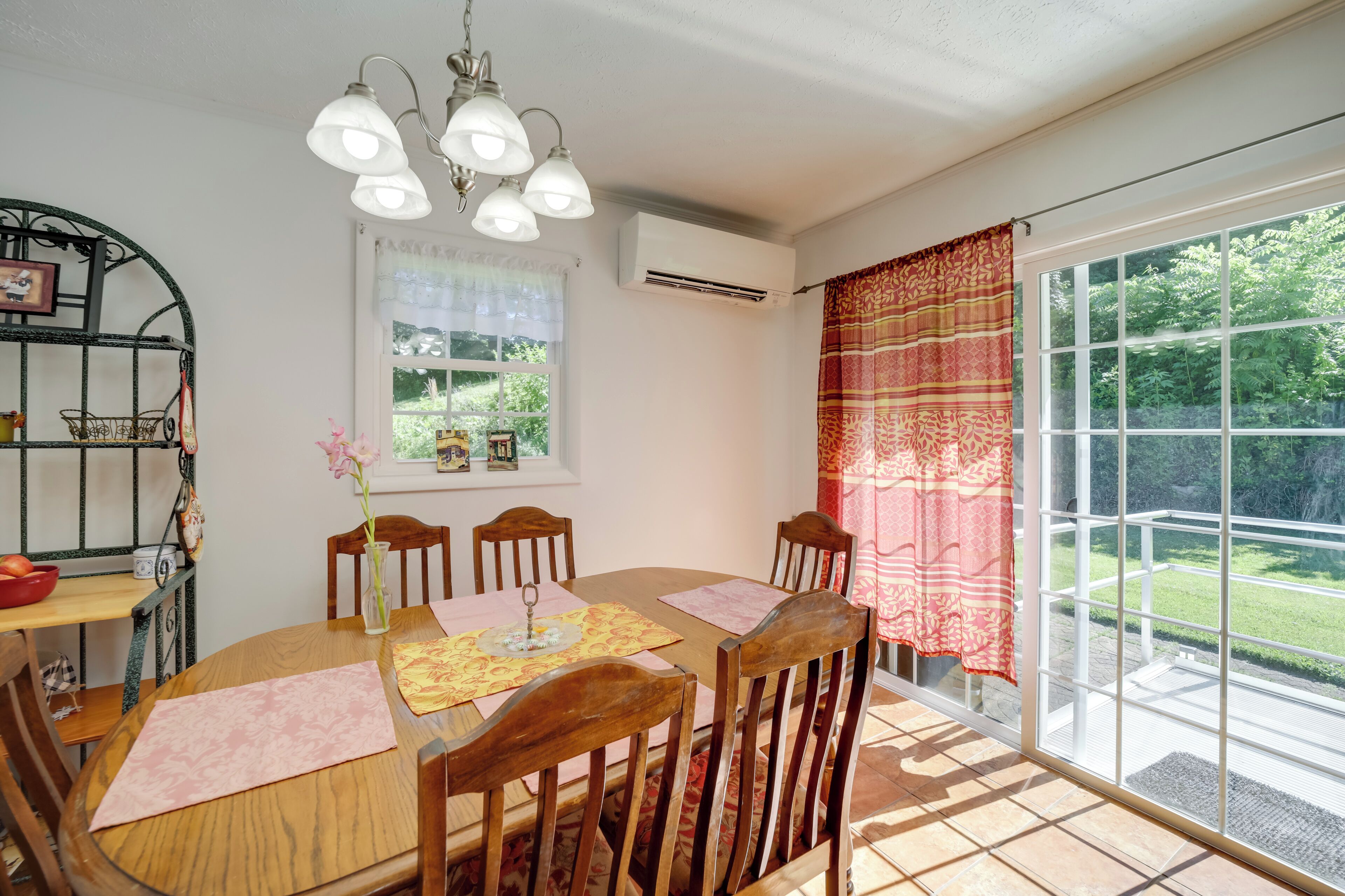 A dining area with a wooden table, chairs, and a chandelier. There is a window with curtains and an air conditioning unit above it.