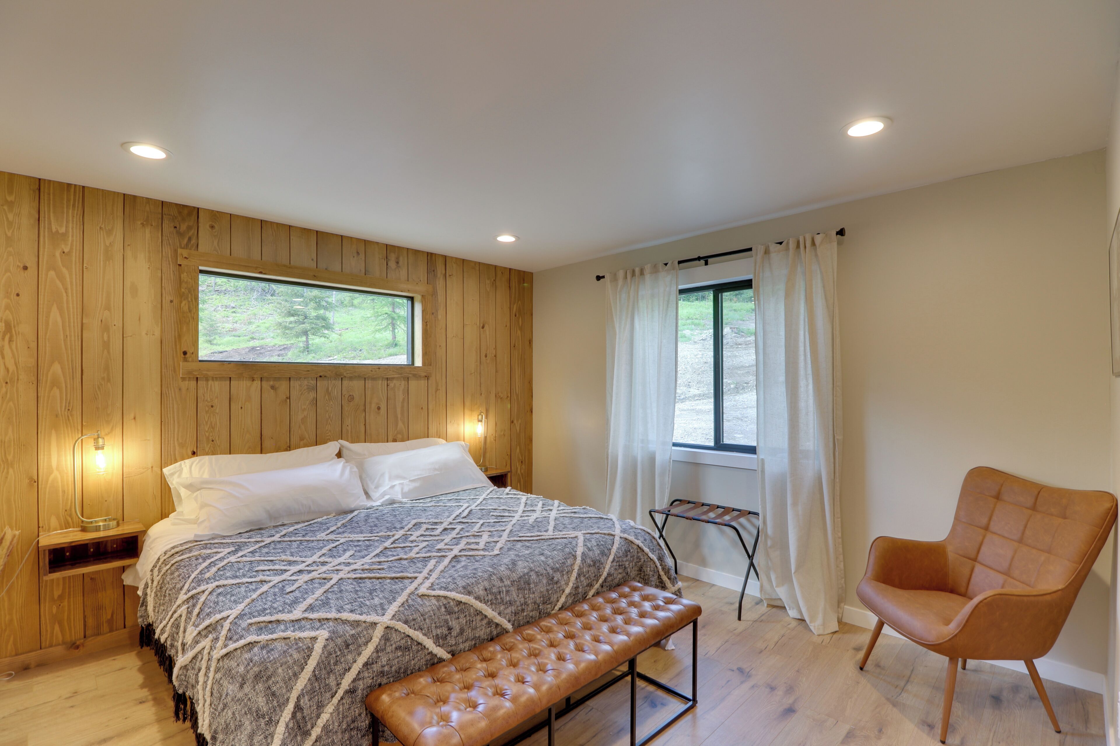 A bedroom with a wooden wall, a large bed with a patterned blanket, a bench, a chair, and a window with curtains.