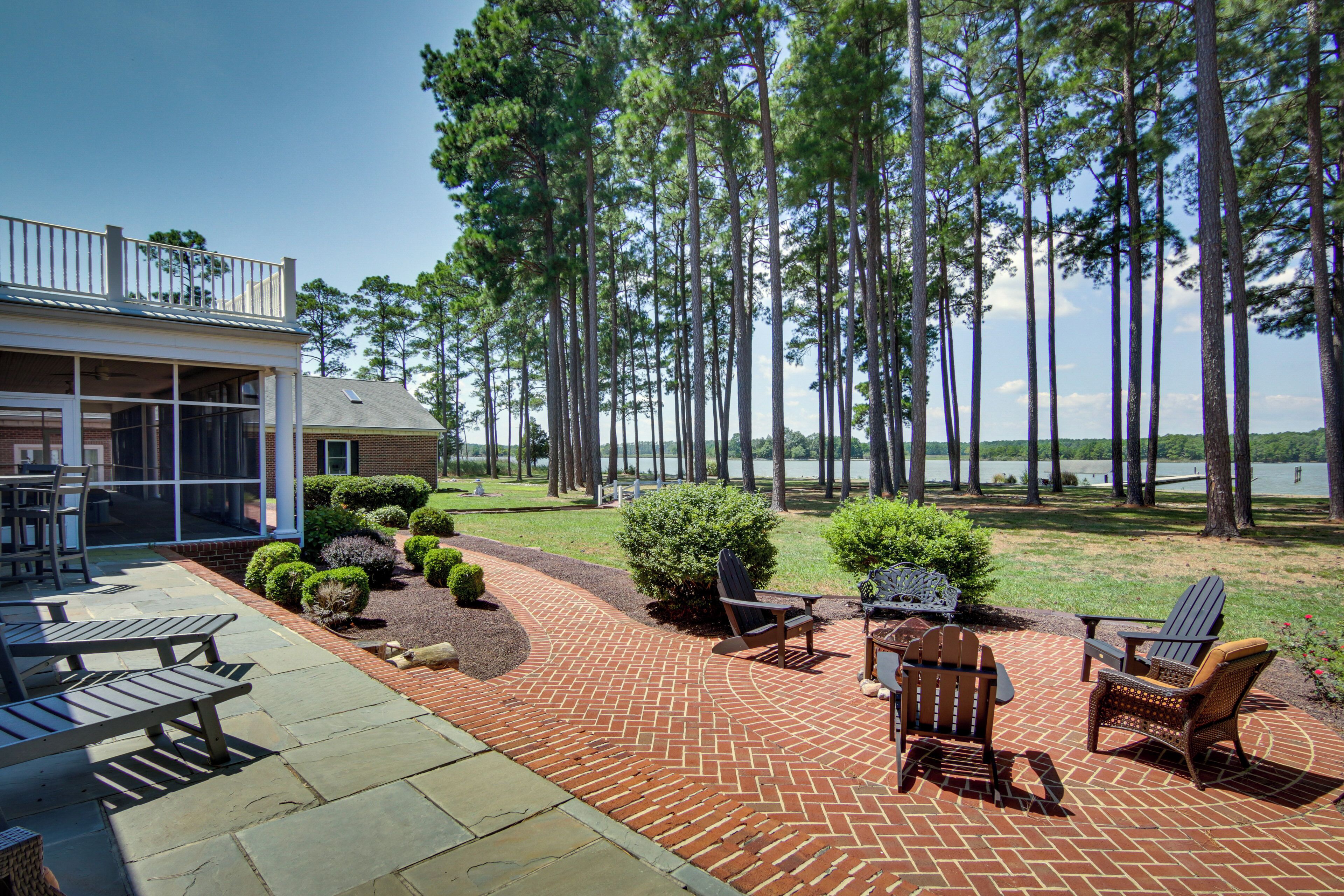 A patio with outdoor furniture, a brick pathway, and a view of a lake surrounded by tall trees.