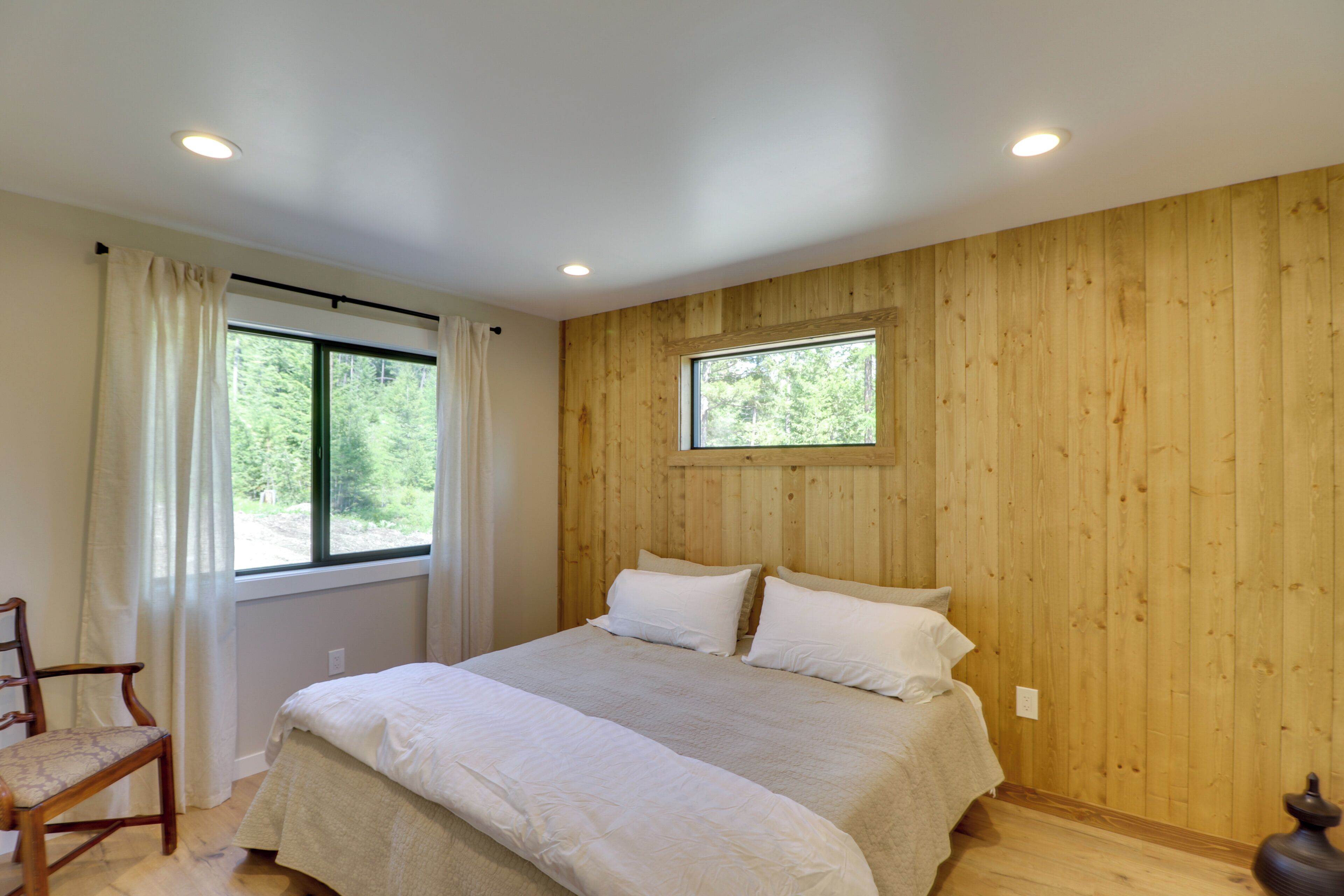 A bedroom with a wooden paneled wall, a large window with curtains, a bed with white bedding, and a wooden chair.