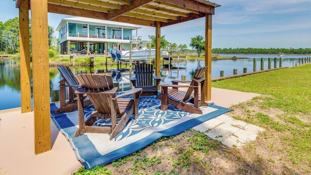 Ein überdachter Außenbereich mit Adirondack-Stühlen und einem blau gemusterten Teppich, mit Blick auf einen See und ein Haus.