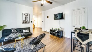 Ein modernes Wohnzimmer mit Deckenventilator, einer schwarzen Couch, einem kleinen runden Tisch mit Pflanze, einem schwarzen Sessel und einem an der Wand befestigten Fernseher.