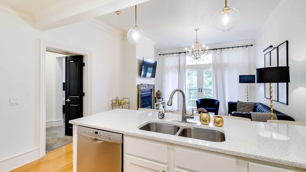 A modern kitchen with a white countertop, stainless steel sink, and a dishwasher. There is a chandelier, a black lamp, and a television mounted on the wall. - Fayetteville Vacation Rental Near UA Campus (Fayetteville)