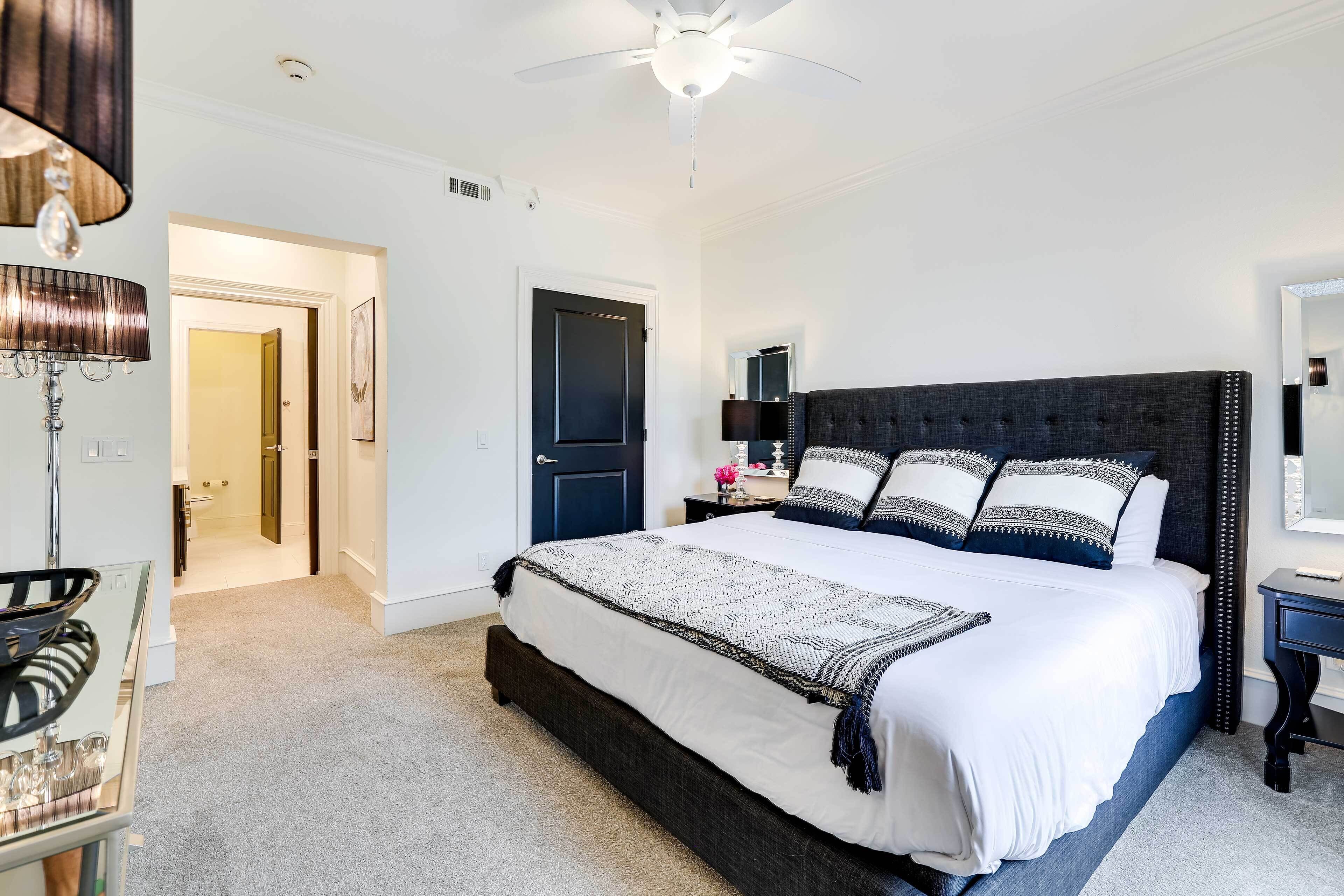 A bedroom with a large bed, a ceiling fan, a bedside table with a lamp, and a door leading to another room.
