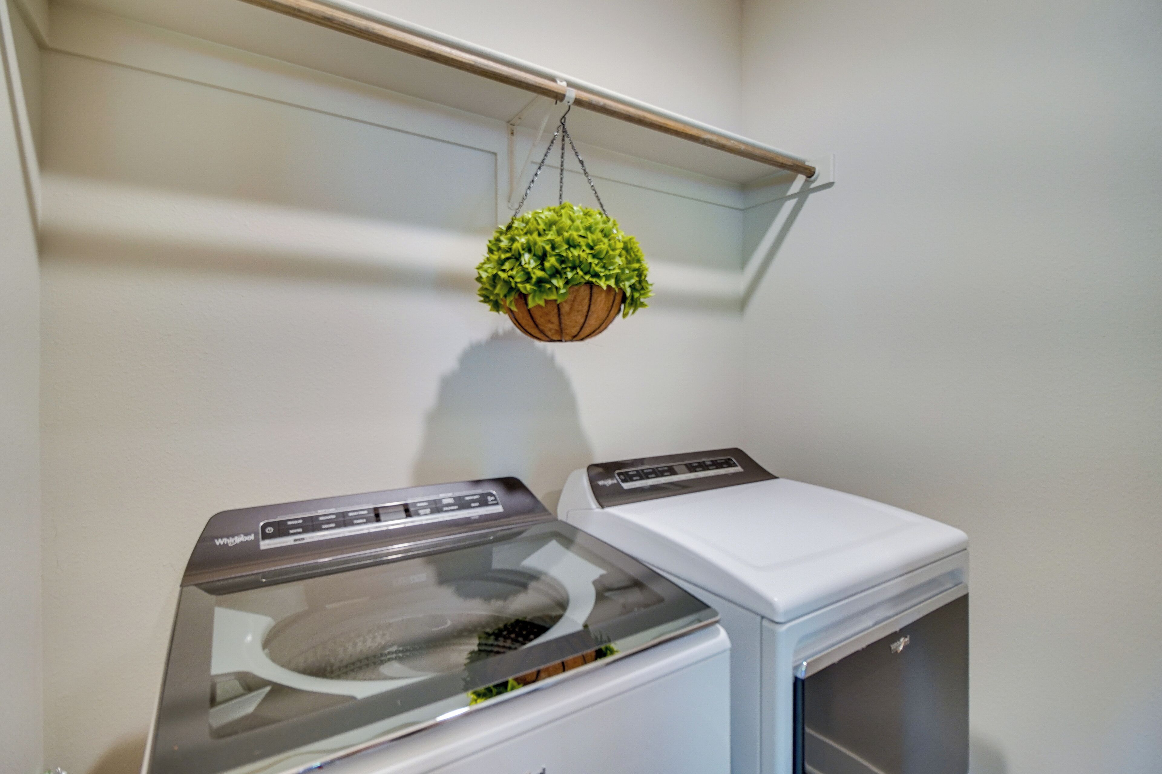 A laundry area with two stacked washers and dryers, a wooden shelf with a hanging plant, and a white wall.