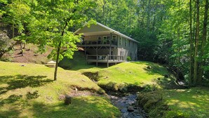Una casa de dos pisos con un balcón, rodeada de un exuberante paisaje verde y un pequeño arroyo.