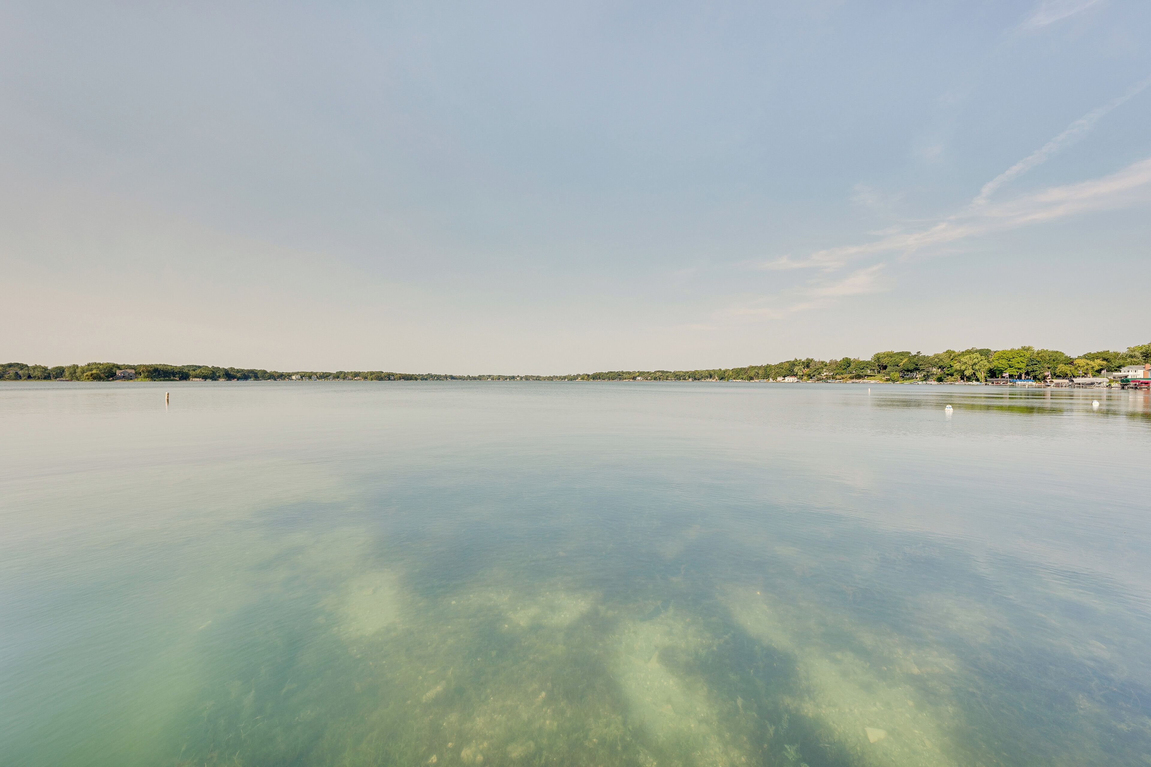 A calm lake with clear water, a distant shoreline with trees and buildings, and a clear sky.
