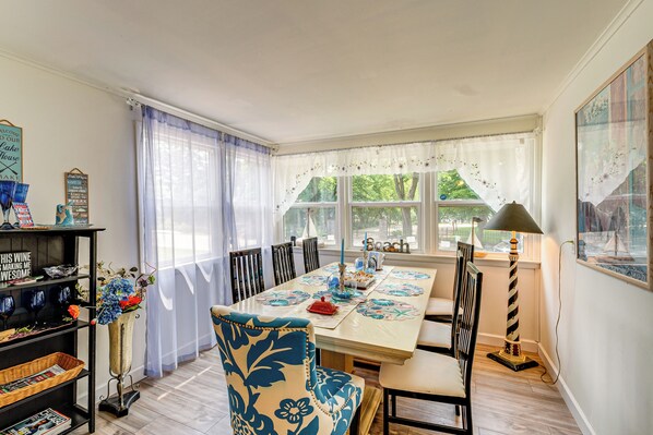 A dining area with a long table set for six, surrounded by chairs. There is a bookshelf to the left, a lamp to the right, and a window with curtains. - Powers Lake Home w/ Patio - 9 Mi to Lake Geneva! (Genoa City)