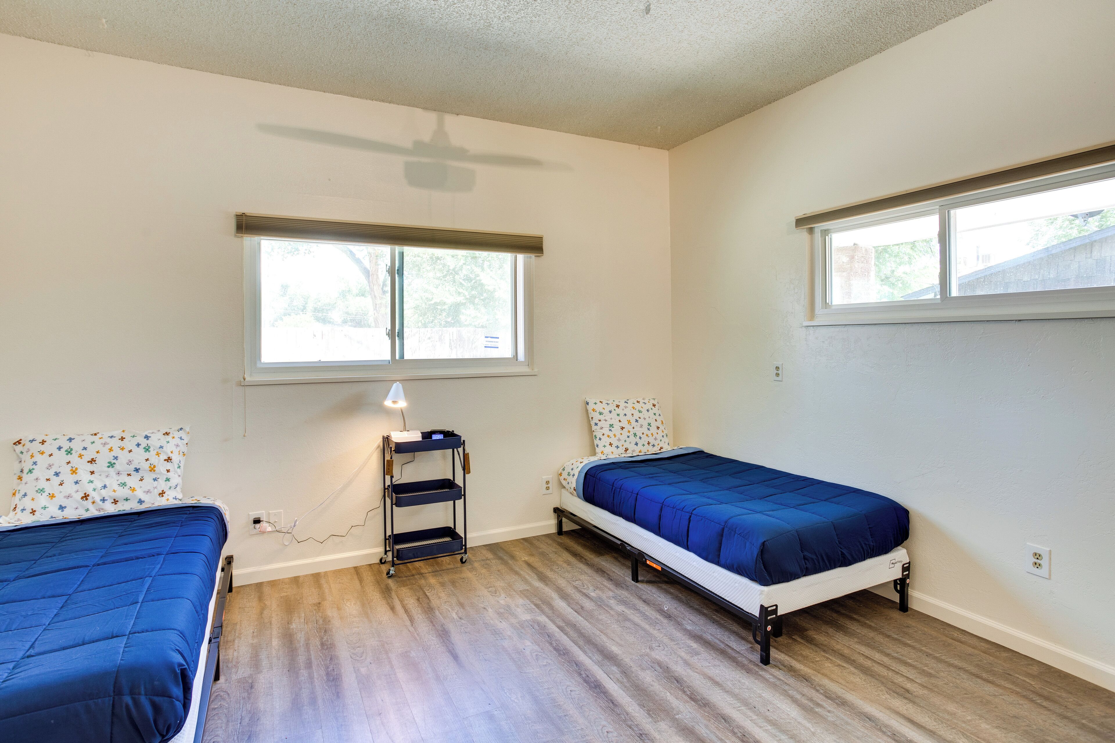 A bedroom with two single beds, a ceiling fan, a window with blinds, and a bedside table with a lamp.