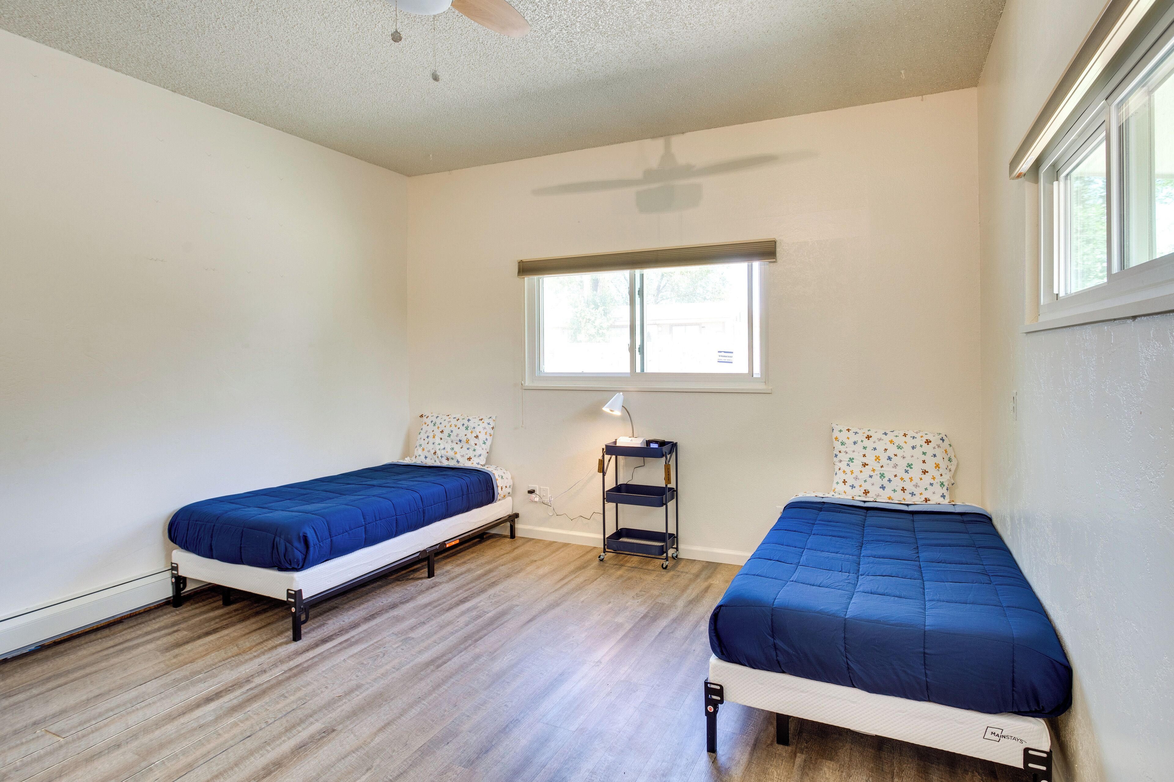 A room with two single beds, a ceiling fan, a window, and a small table with a lamp.