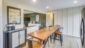 Een moderne eethoek met een lange houten tafel, zwarte metalen stoelen, een koffieapparaat en een menukaart in een lijst aan de muur.