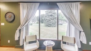 Een kamer met twee witte stoelen, een klein rond tafeltje, een klok aan de muur en een raam met gordijnen.