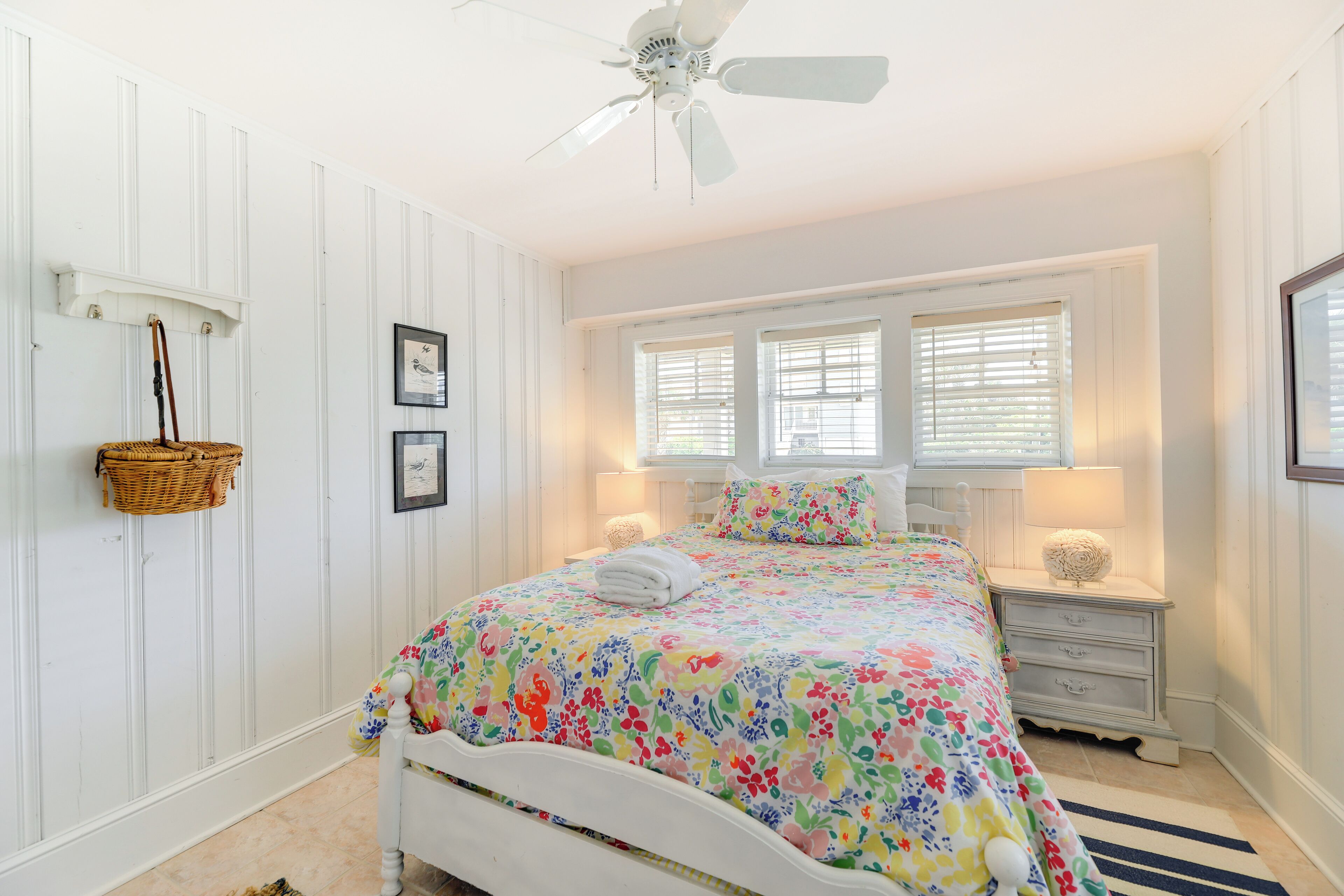 A bedroom with a white bed, floral bedding, a ceiling fan, and two windows with blinds.