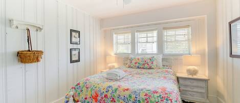 A bedroom with a white bed, floral bedding, a ceiling fan, and two windows with blinds.