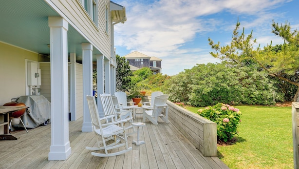 Une terrasse en bois, agrémentée de chaises berçantes blanches et d’un barbecue, qui offre une vue sur une pelouse et un jardin bien entretenus.