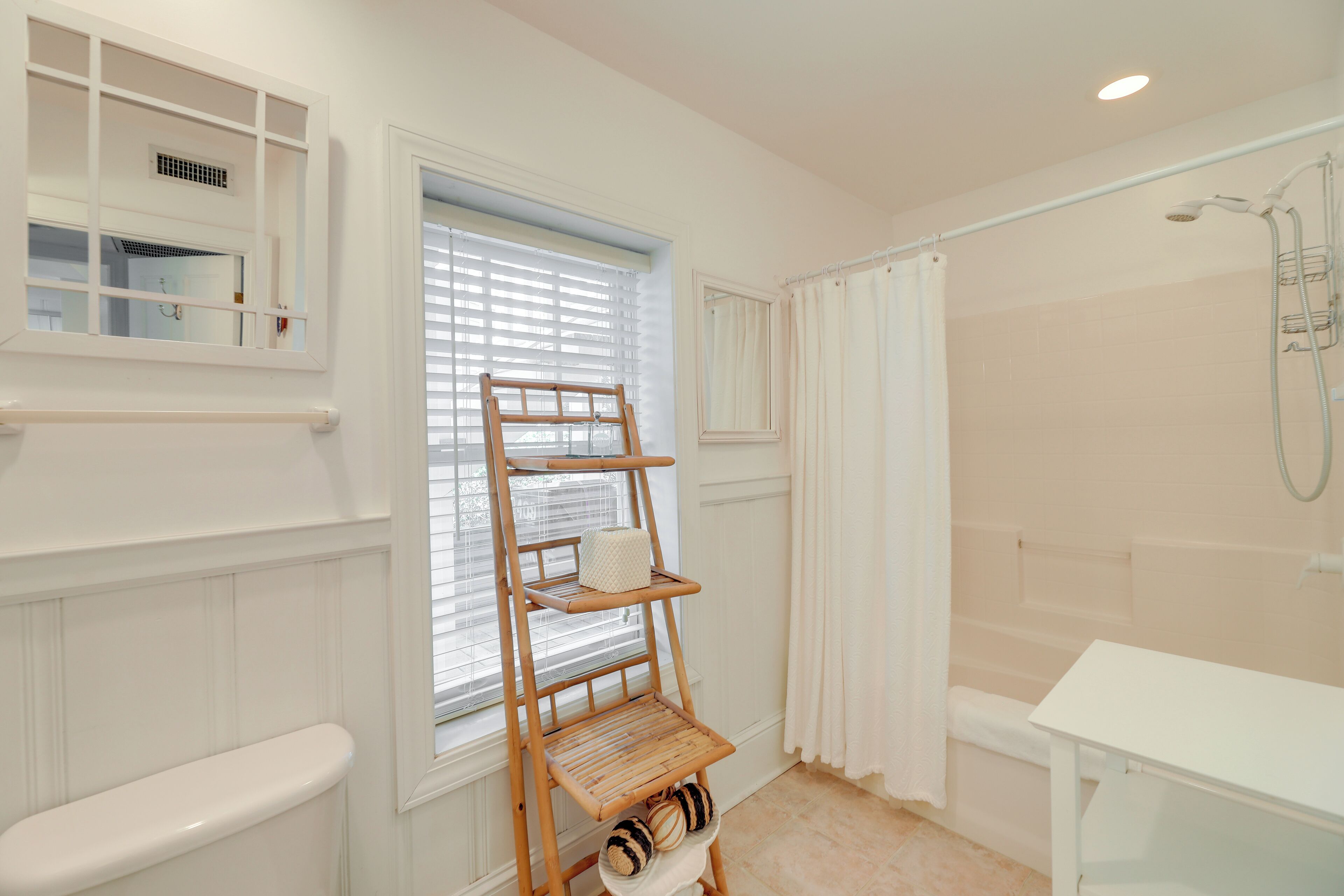 Un baño con inodoro, una ventana con persianas, un estante de madera con toallas, una zona de ducha y un lavabo blanco.