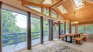 A spacious room with large windows, a wooden ceiling, and a view of a forested area.