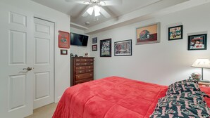A bedroom with a red bedspread, a ceiling fan, a dresser, and several framed pictures on the wall.