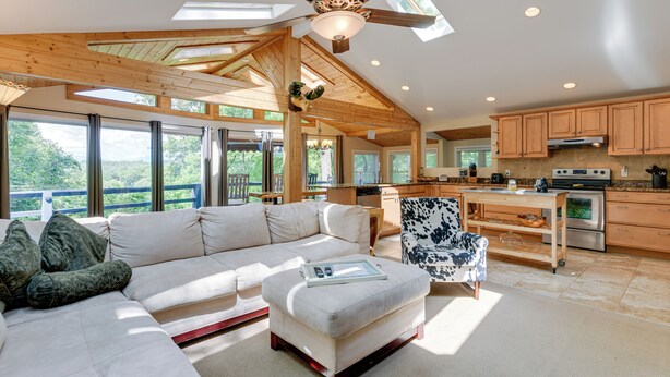 A spacious living room with a high ceiling, large windows, and a modern kitchen with wooden cabinets and stainless steel appliances.