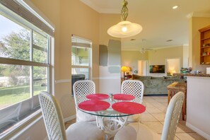 A dining area with a glass table surrounded by white wicker chairs, a kitchen in the background, and a view of the outdoors. - Waterfront Lake Placid Home w/ Yard & Boat Launch! (Lake Placid)