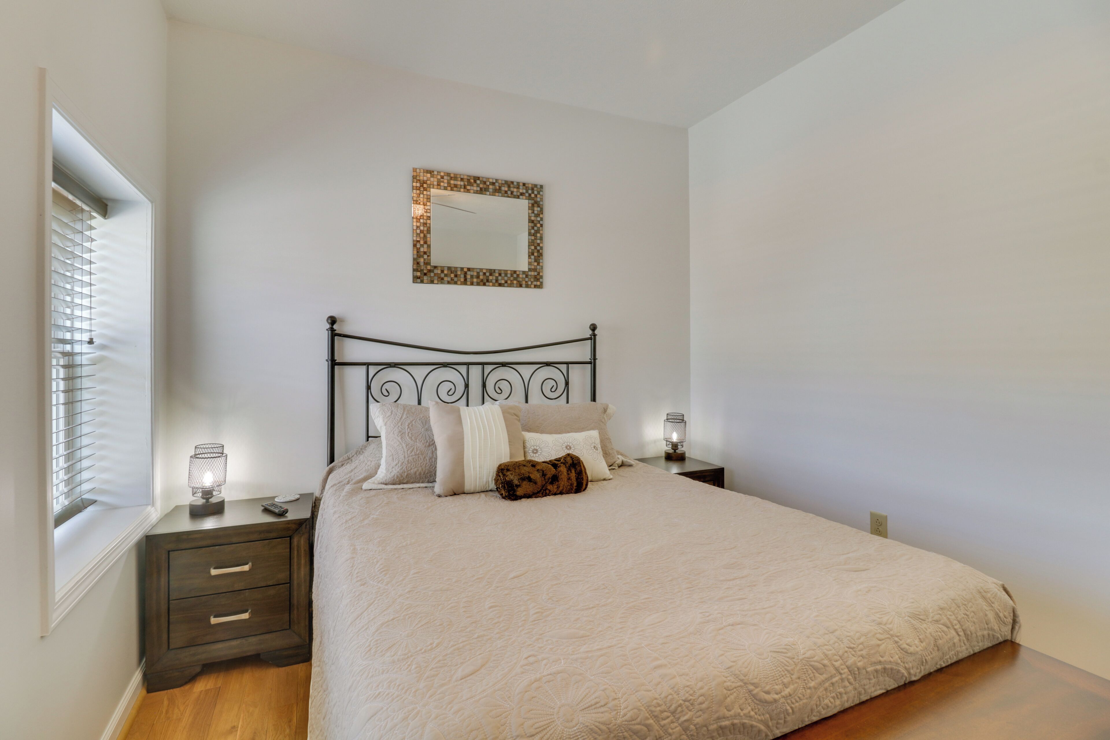 A bedroom with a bed, two bedside tables, a nightstand, a mirror, and a window with blinds.