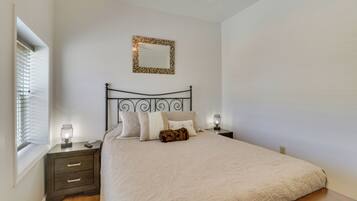 A bedroom with a bed, two bedside tables, a nightstand, a mirror, and a window with blinds.