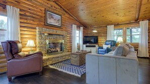 A log cabin interior with a stone fireplace, leather armchair, white sofa, and a television. - Cozy Brookville Home w/ Fire Pit & Deck! (Brookville)