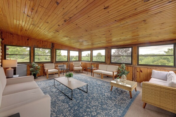Ein geräumiger Raum mit Holzwänden und -decke, großen Fenstern mit Blick auf Grünflächen sowie einer Mischung an Sitzgelegenheiten, darunter ein Sofa, Stühle und ein kleiner Tisch.