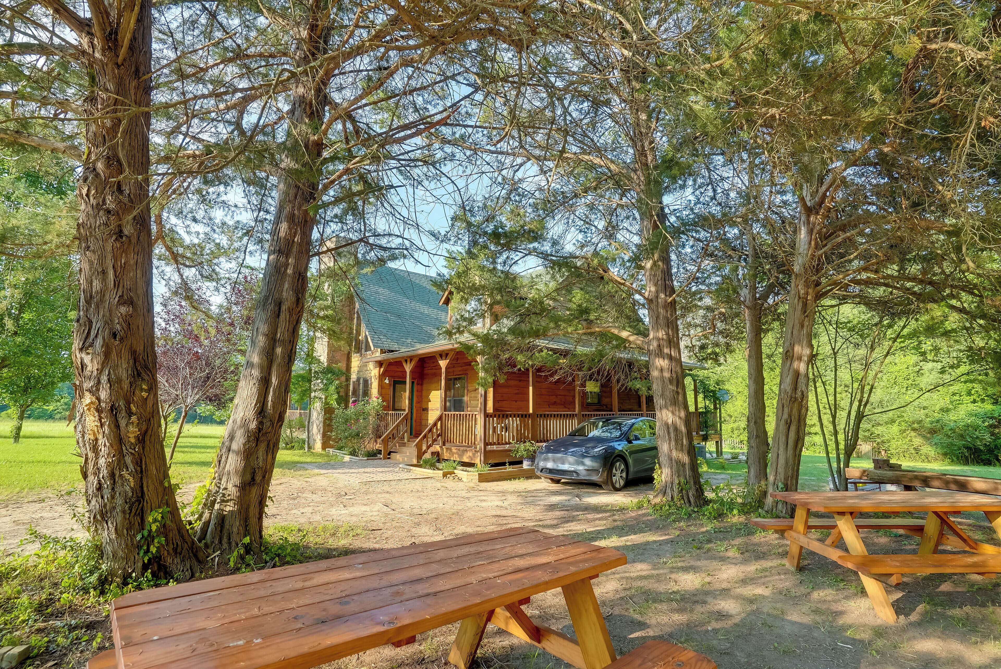 A rustic cabin with a covered porch, a car parked in front, and picnic tables in a wooded area.