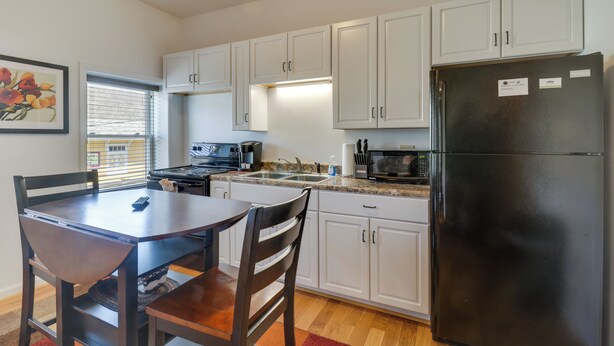 Una cocina compacta con armarios blancos, un refrigerador negro, una mesa de comedor con sillas y una ventana con persianas.