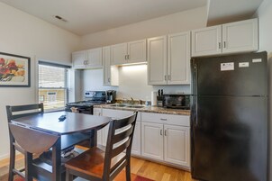 Una cocina compacta con armarios blancos, un refrigerador negro, una mesa de comedor con sillas y una ventana con persianas.
