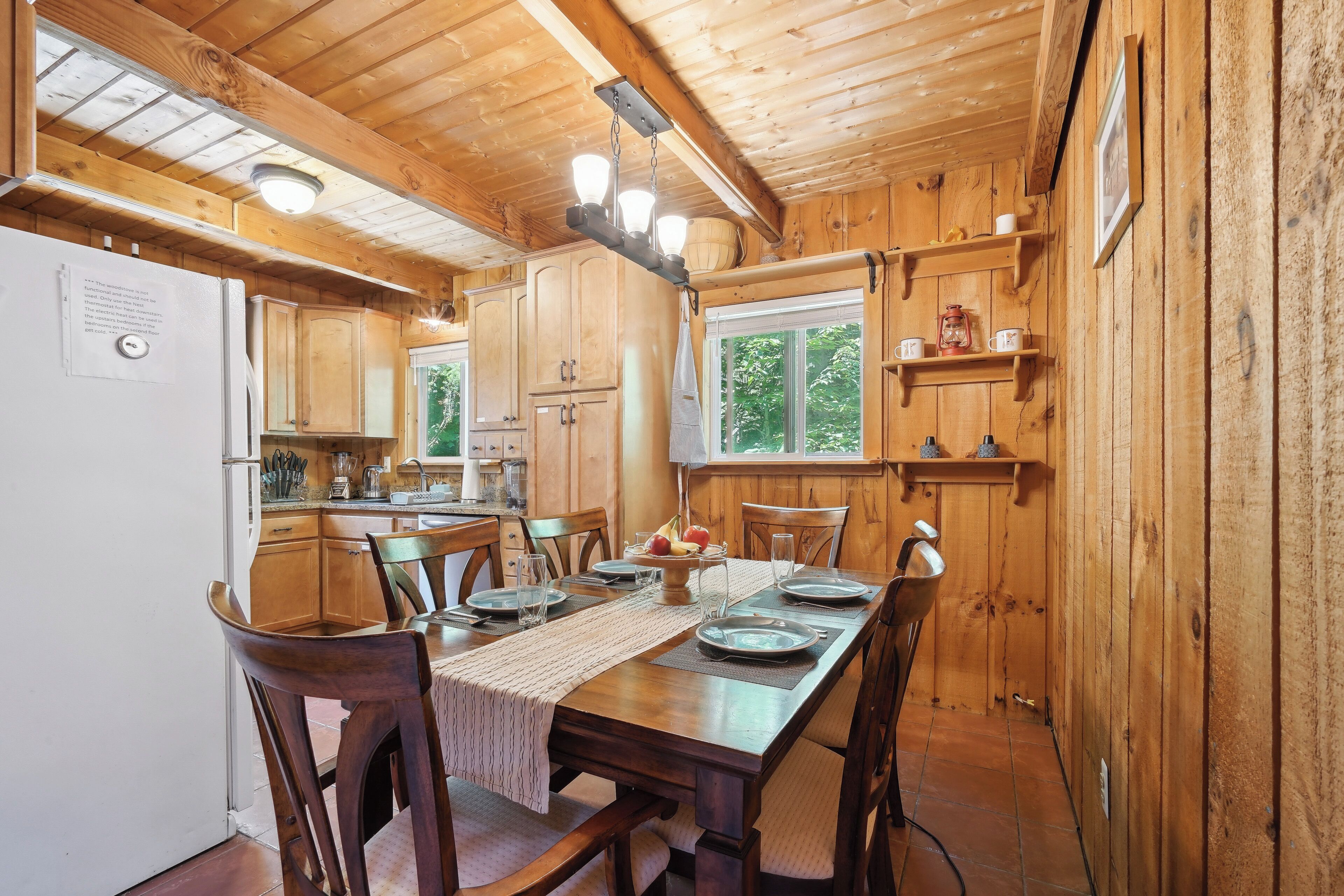 A wooden cabin interior with a dining area, kitchen, and a window overlooking greenery.