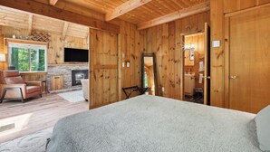 Interior de una cabaña de madera con chimenea, televisor, cama y baño visible a través de una puerta abierta.