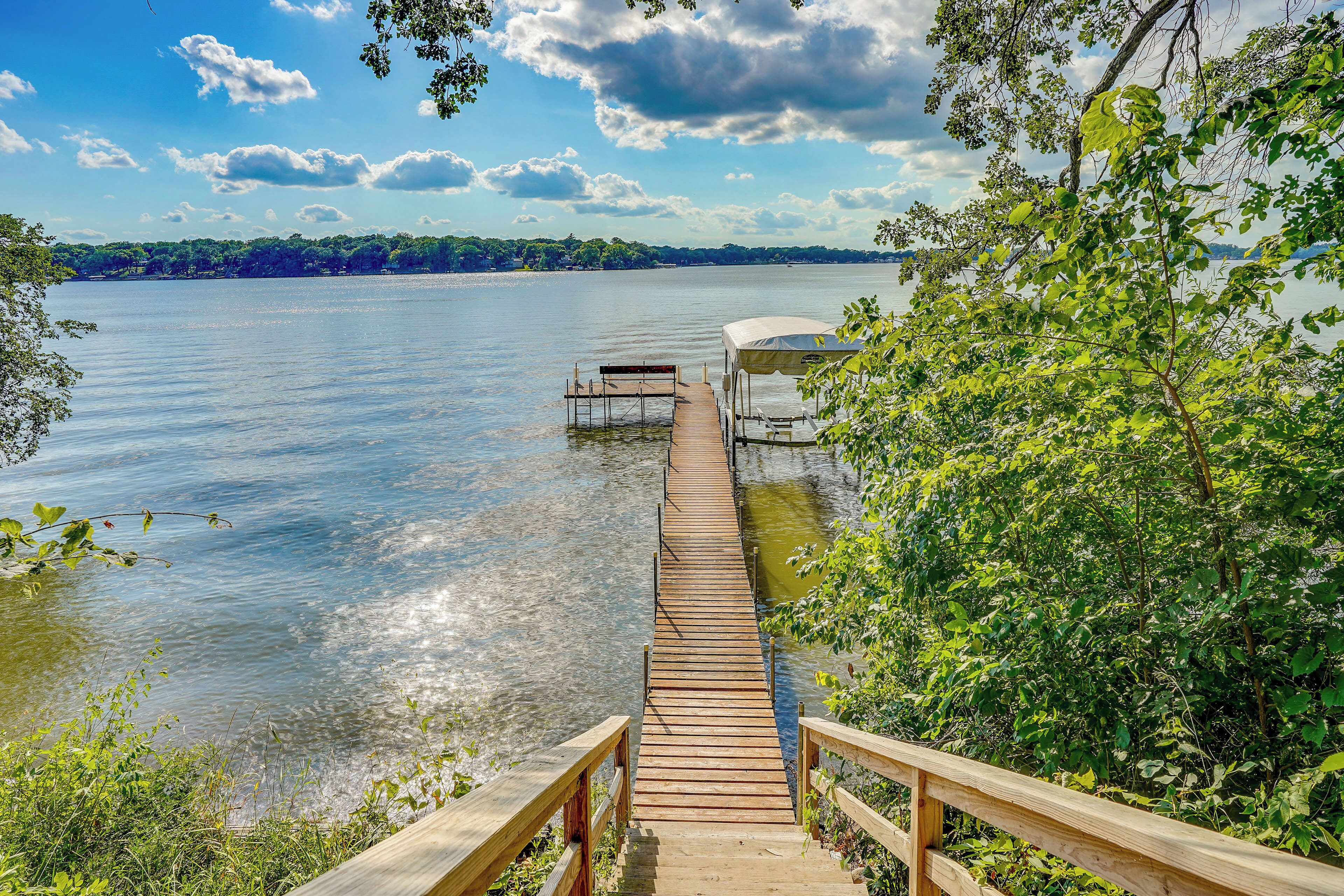 Un muelle de madera que lleva a una zona cubierta para el lanzamiento de embarcaciones junto a un lago tranquilo, rodeado de abundante vegetación.