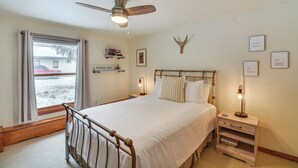 Ett sovrum med en säng, sängbord, en takfläkt, ett fönster med gardiner, en vägghylla med böcker och en dekorativ hjorthuvudtrofé.