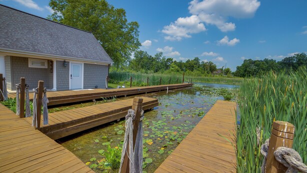A wooden dock leading to a house with a gray roof, surrounded by a pond with lily pads and tall grass. - Jackson Lakefront Getaway w/ Balcony, Dock Access (Jackson)