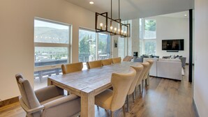 A spacious dining area with a large wooden table and comfortable chairs, large windows offering a view of the outdoors, and a modern kitchen in the background. - Avon Vacation Rental w/ Hot Tub & Mountain Views! (Avon)