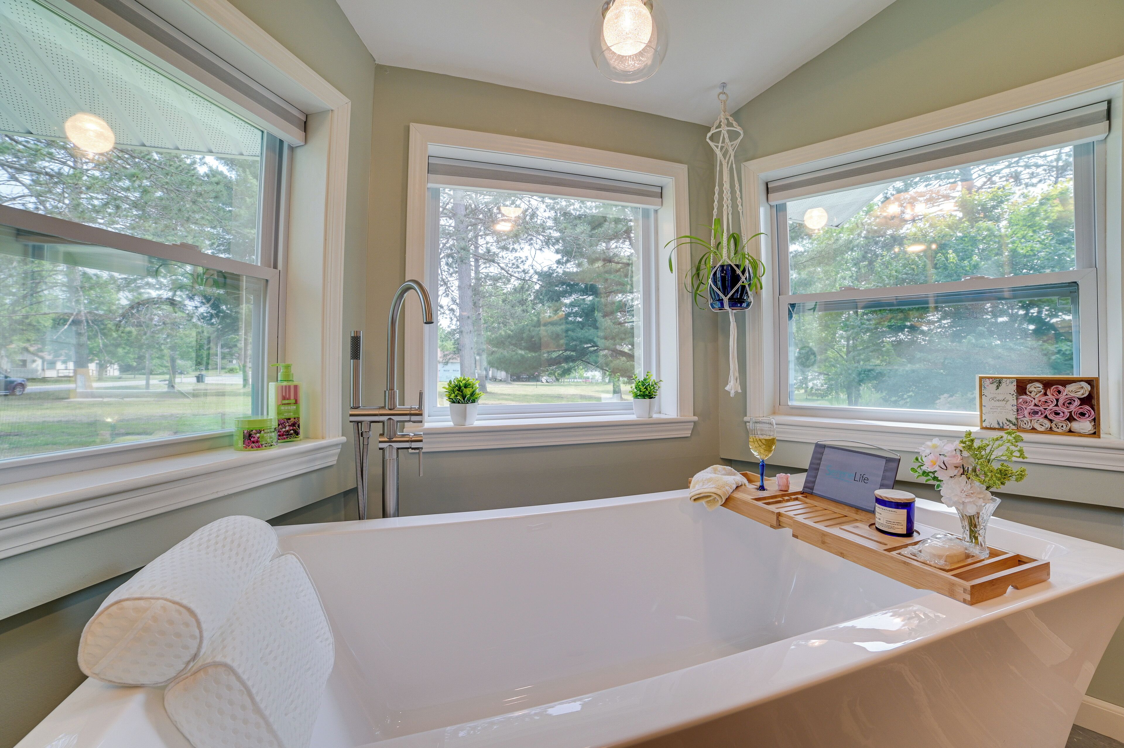 A modern bathroom with a large bathtub, two windows with views of greenery, and a laptop on the edge of the tub.