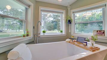 A modern bathroom with a large bathtub, two windows with views of greenery, and a laptop on the edge of the tub.