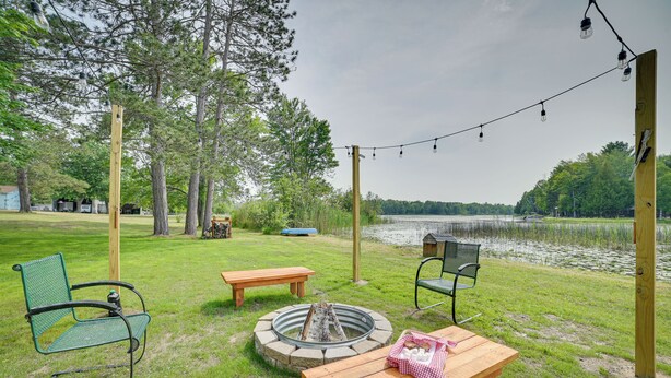 A lakeside patio with a fire pit, picnic table, and chairs, surrounded by trees and a view of the lake. - Northern Michigan Vacation Rental Near Trails! (Atlanta)